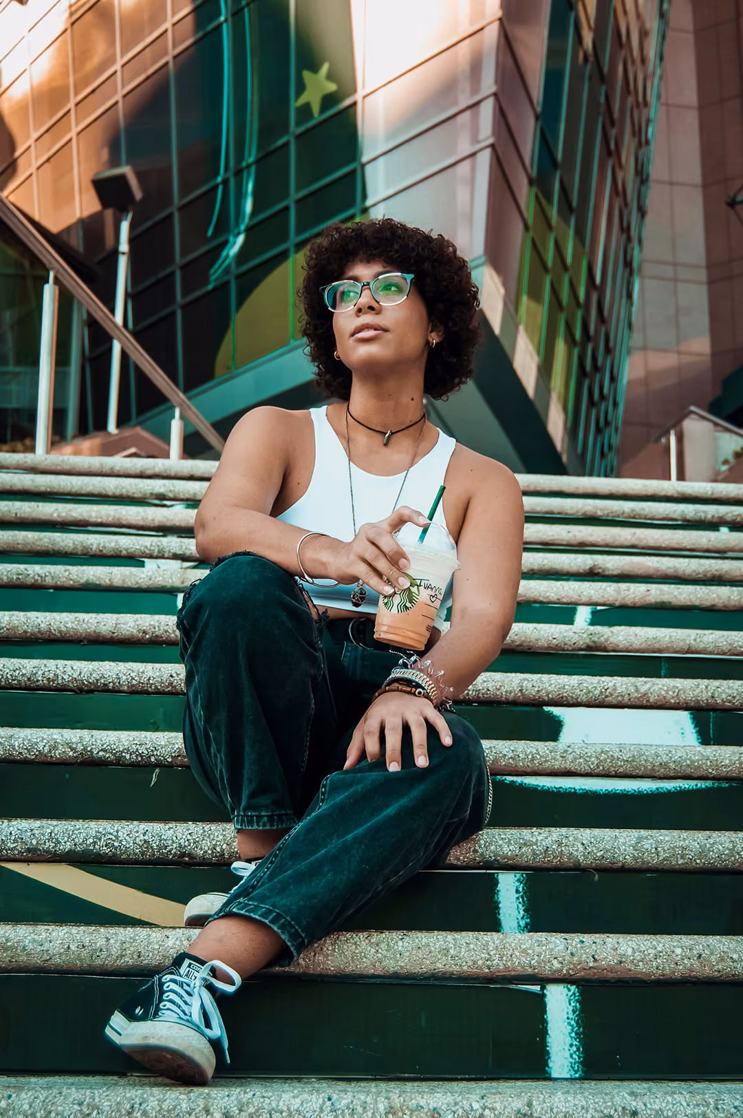 Young woman with curly hair and glasses sitting on stairs holding a Starbucks iced drink.