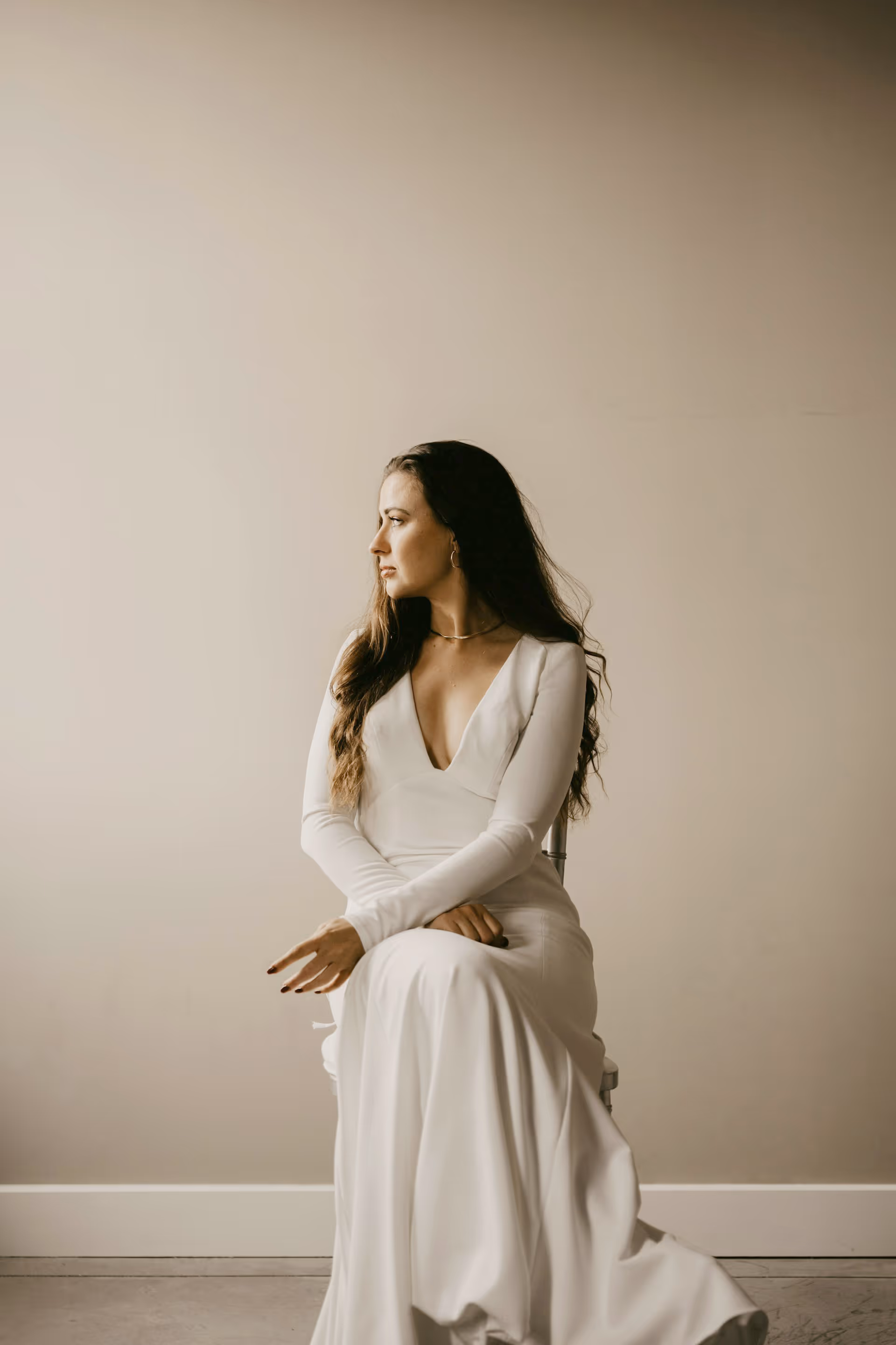 Woman with long brown hair sitting on a chair wearing a white long-sleeved dress looking to the side.