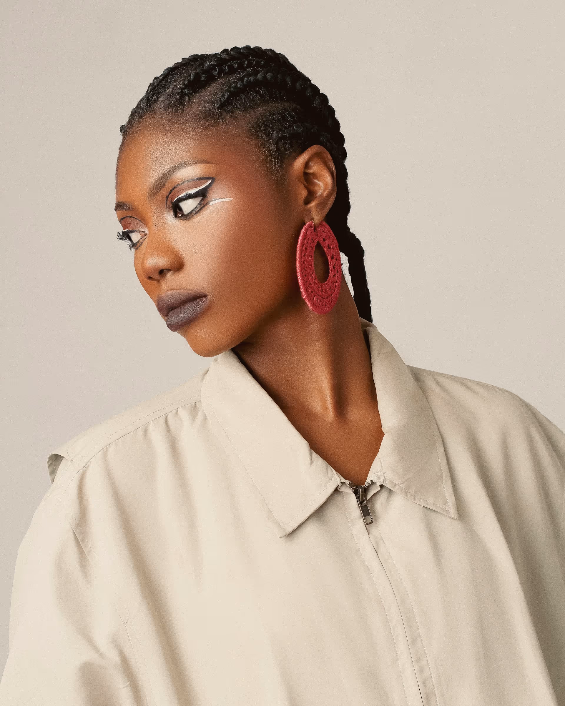 Profile of a woman with braided hair wearing bold black and white eye makeup and large red circular earrings, dressed in a beige jacket.