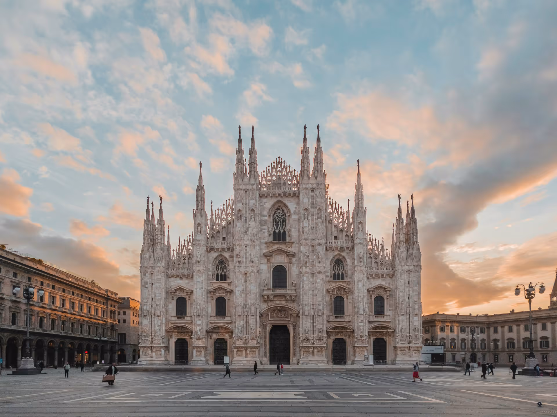 Front view of Milan Cathedral with intricate Gothic architecture at sunset and people walking nearby.