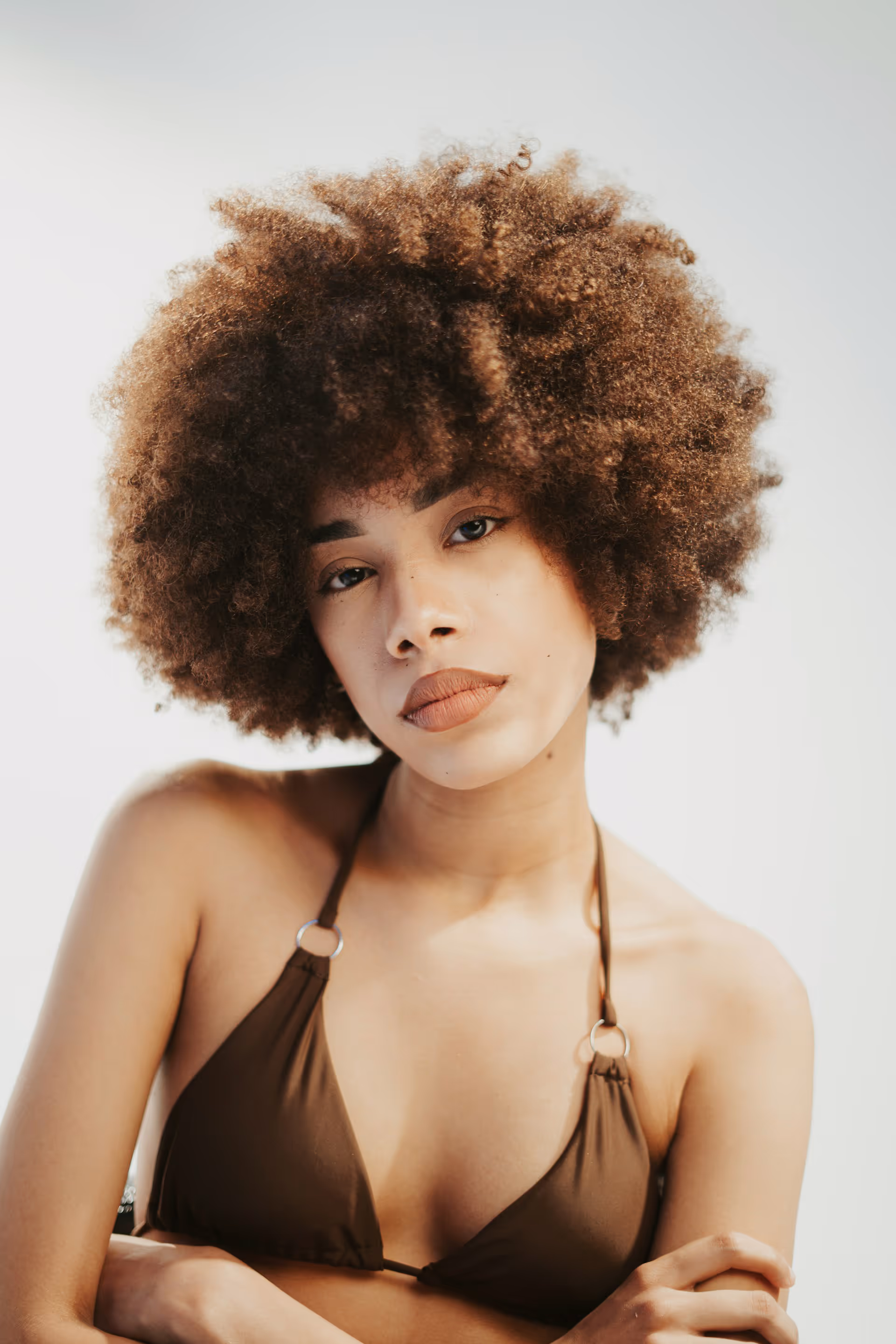 Portrait of a woman with natural curly hair wearing a brown halter top against a light background.