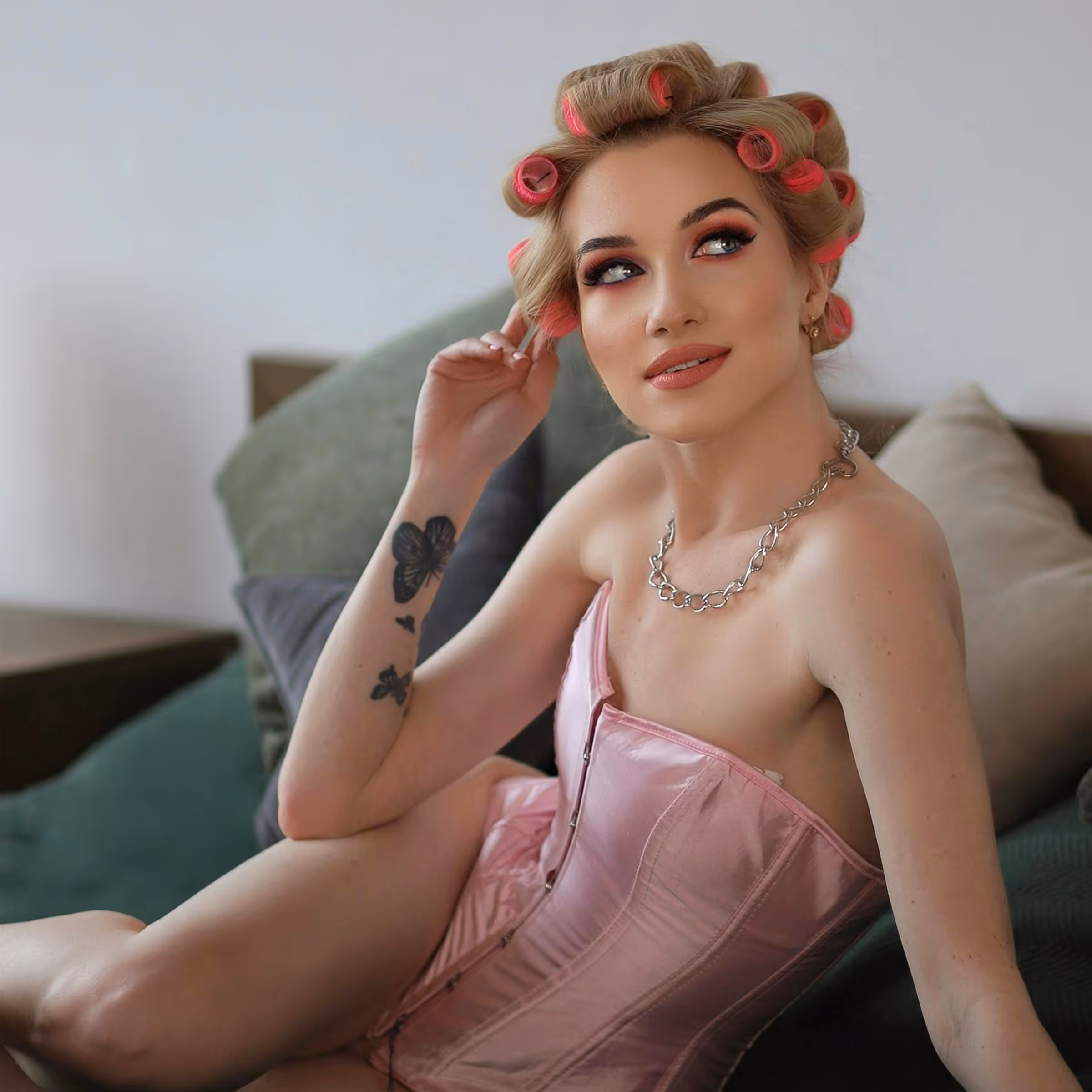 Woman with pink hair rollers wearing a pink corset and a silver chain necklace, sitting on a couch with butterfly tattoos on her arm.