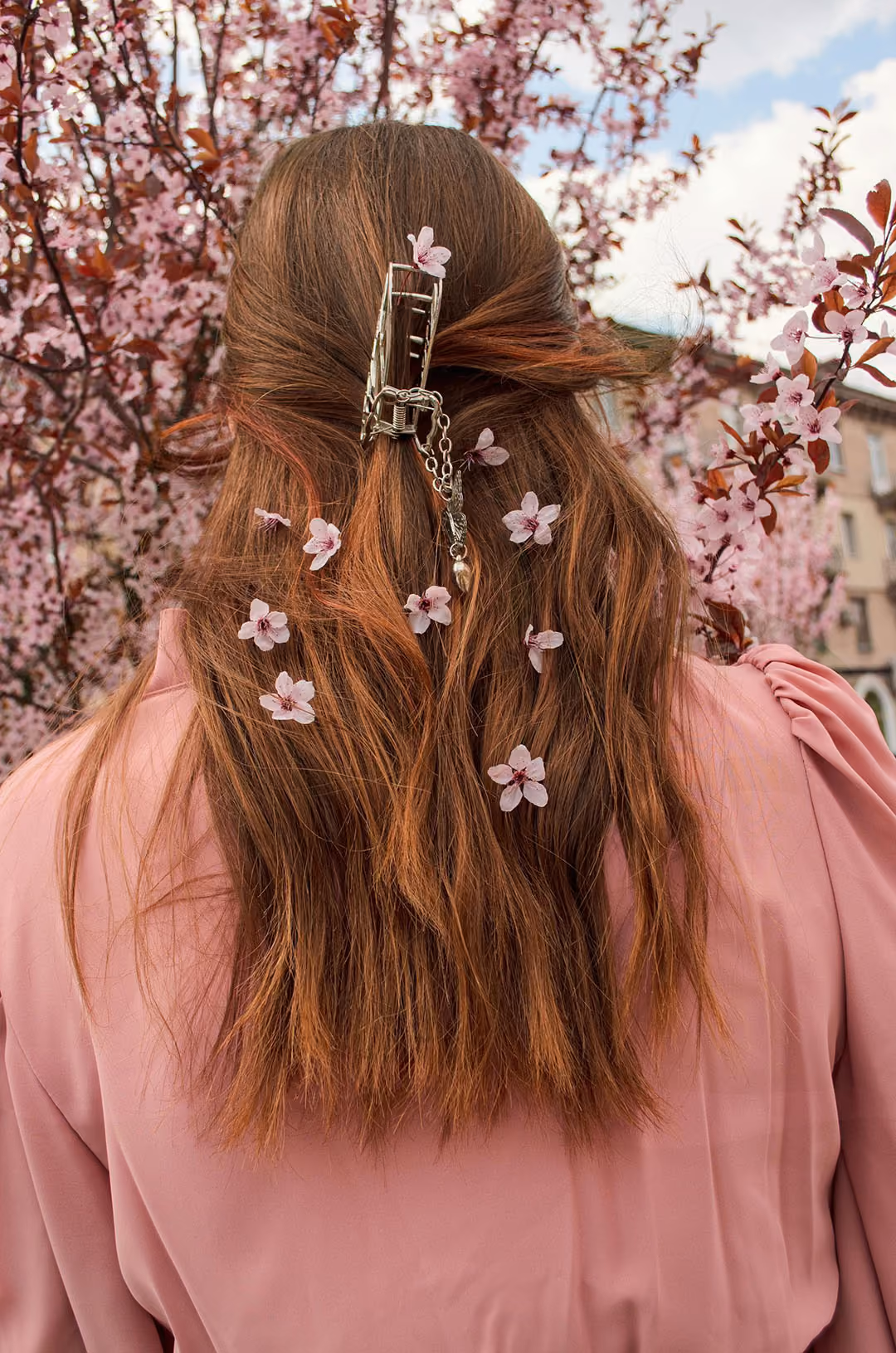 Back view of a person with long reddish hair adorned with pink cherry blossom flowers and held by a silver hair clip, wearing a pink top, standing in front of a cherry blossom tree.