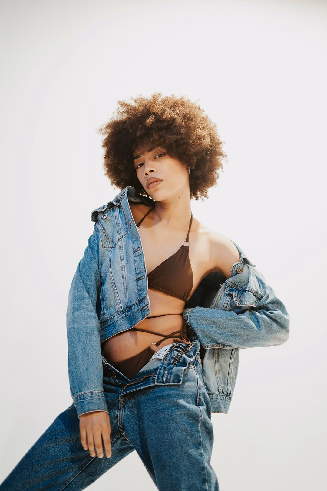 Young woman with curly afro wearing a brown bikini top under a denim jacket and blue jeans against a plain light background.