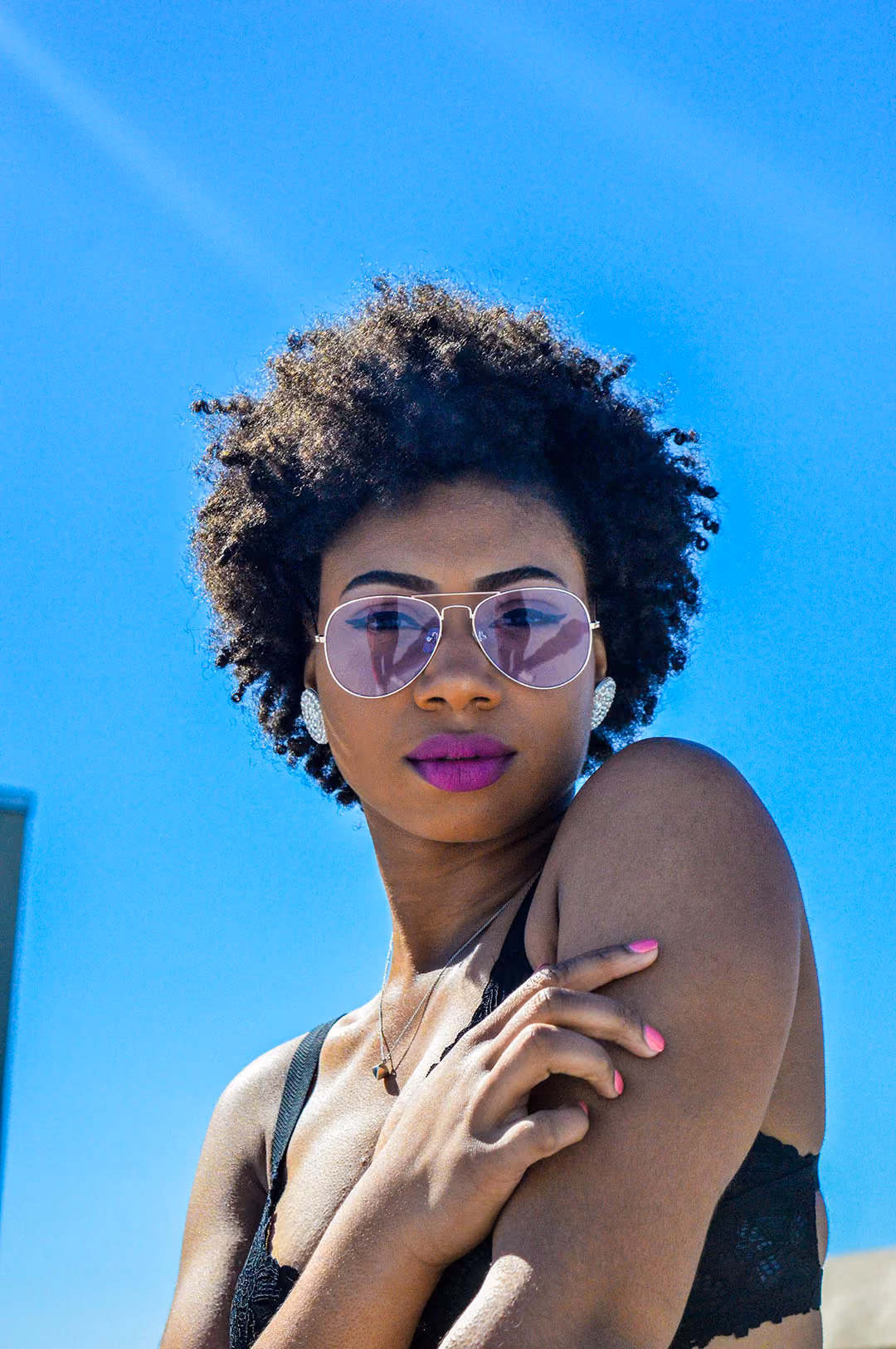 Woman with curly hair wearing purple sunglasses, bright pink lipstick, and a black lace top against a clear blue sky.