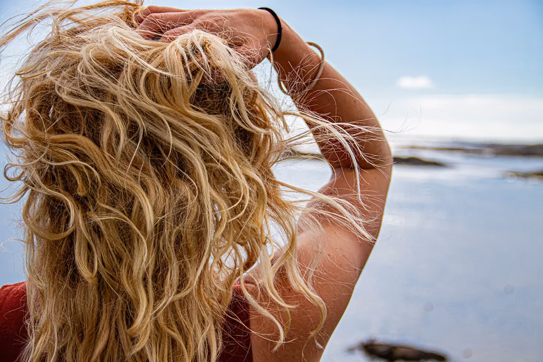 Person with long, curly blonde hair holding one arm up, facing a calm body of water under a clear sky.