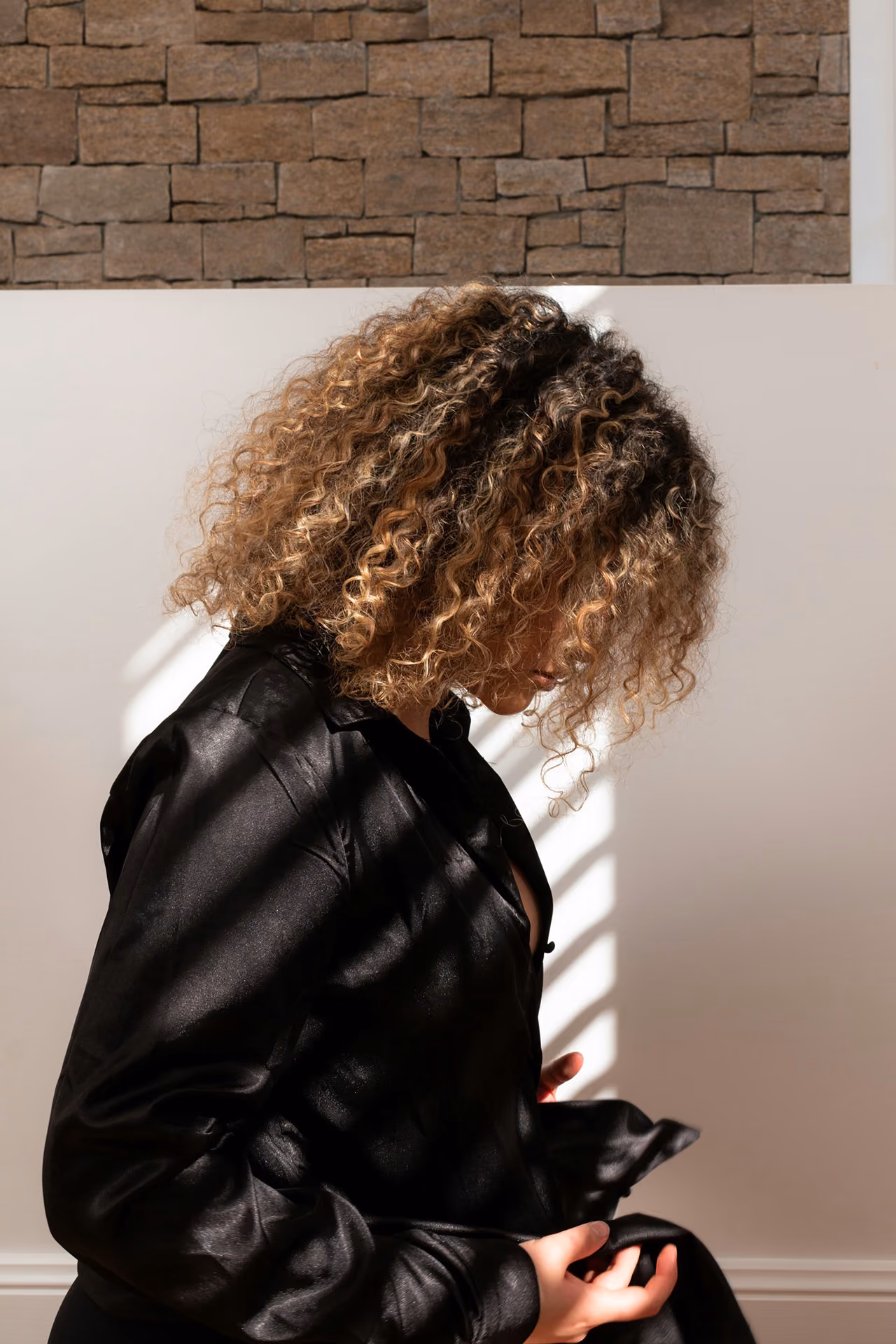 Person with curly hair wearing a black satin shirt, looking down with sunlight casting shadows.