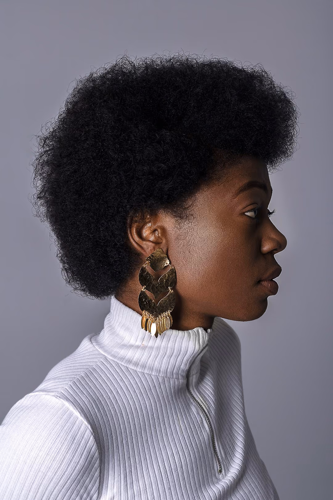 Profile of a person with natural afro hair wearing large gold earrings and a white ribbed turtleneck sweater.