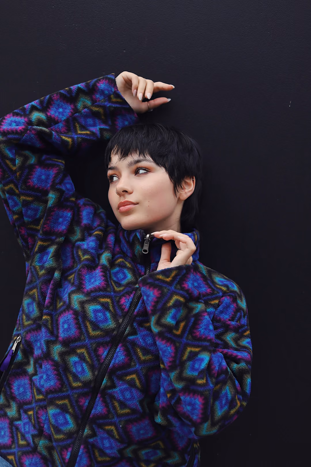 Young person with short black hair wearing a colorful patterned fleece jacket, posing against a black background.