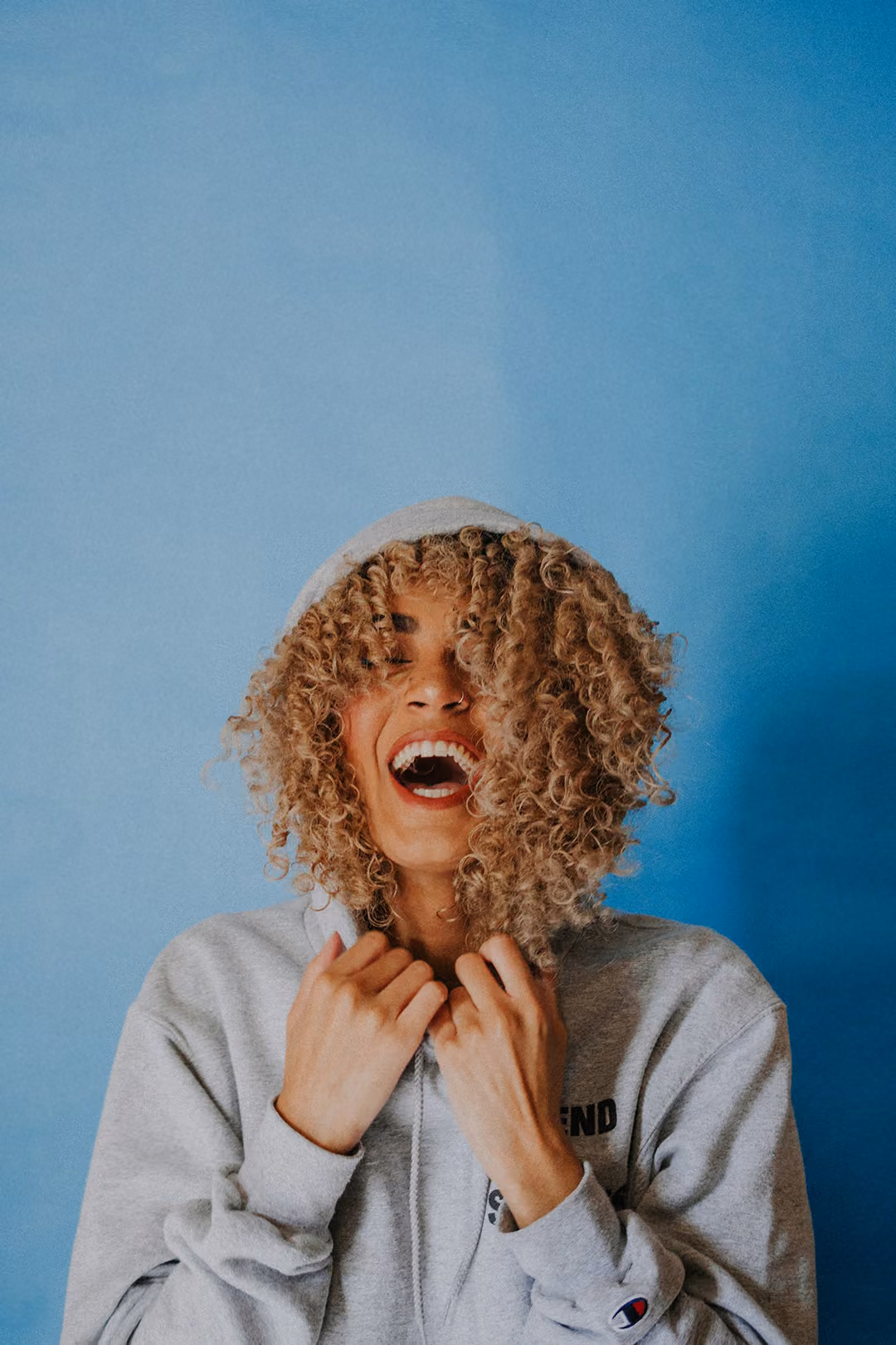 Smiling person with curly hair wearing a gray hoodie against a blue background.