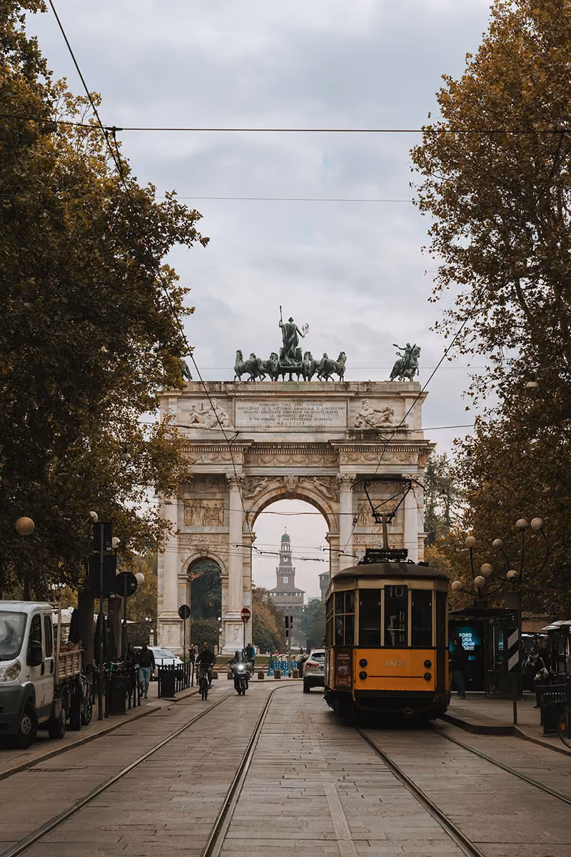 Yellow tram on tracks passing under Arco della Pace with Castle Sforzesco visible in the background, Milan.