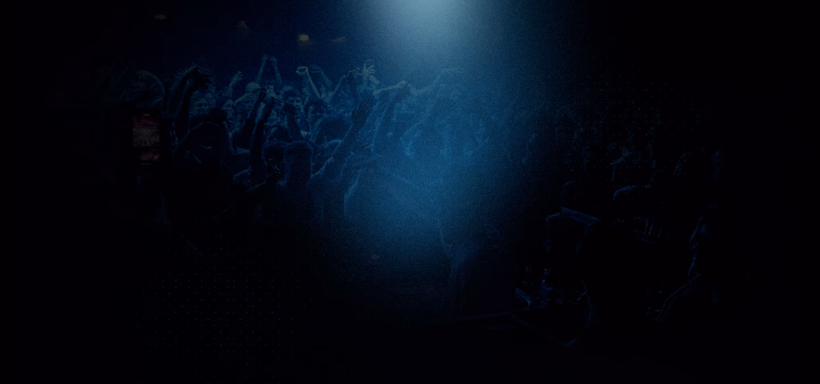 Dimly lit crowd raising hands and recording with phones at a concert or event under a blue spotlight.