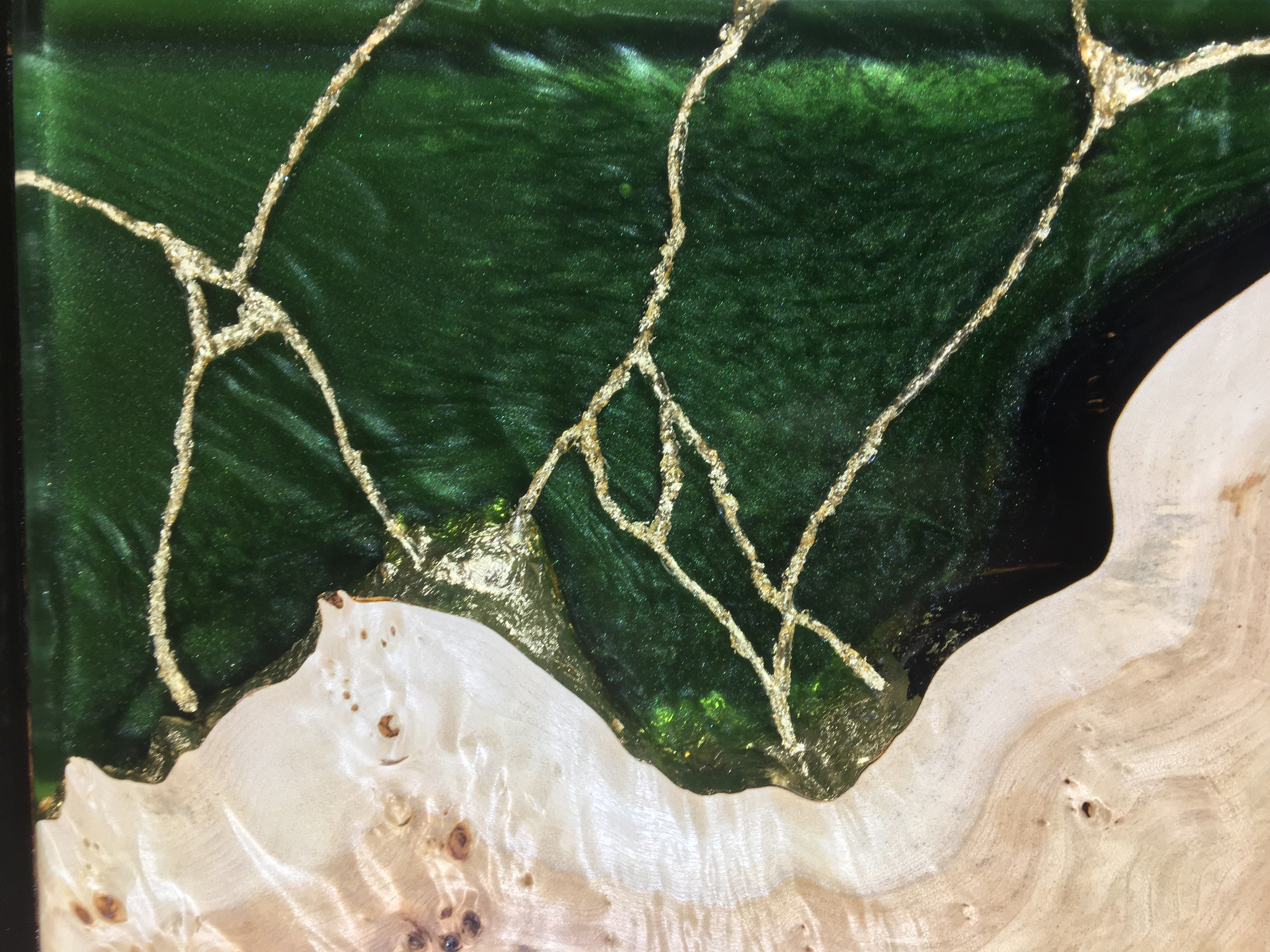 Détail d'une surface en bois clair combinée à une résine époxy verte avec des fissures comblées de poudre dorée.