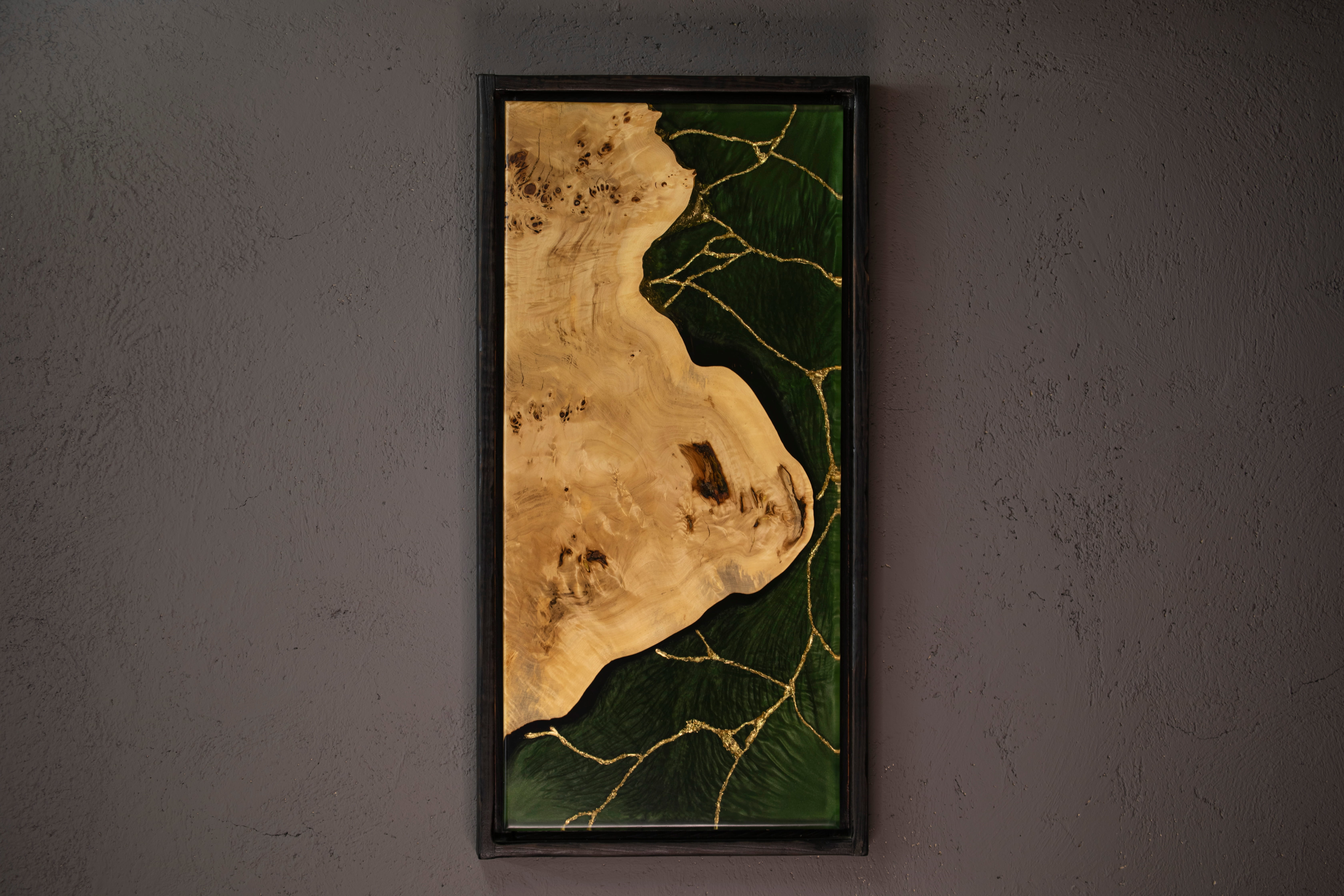 Tableau en bois naturel avec résine verte et fissures dorées, encadré en noir sur un mur gris.