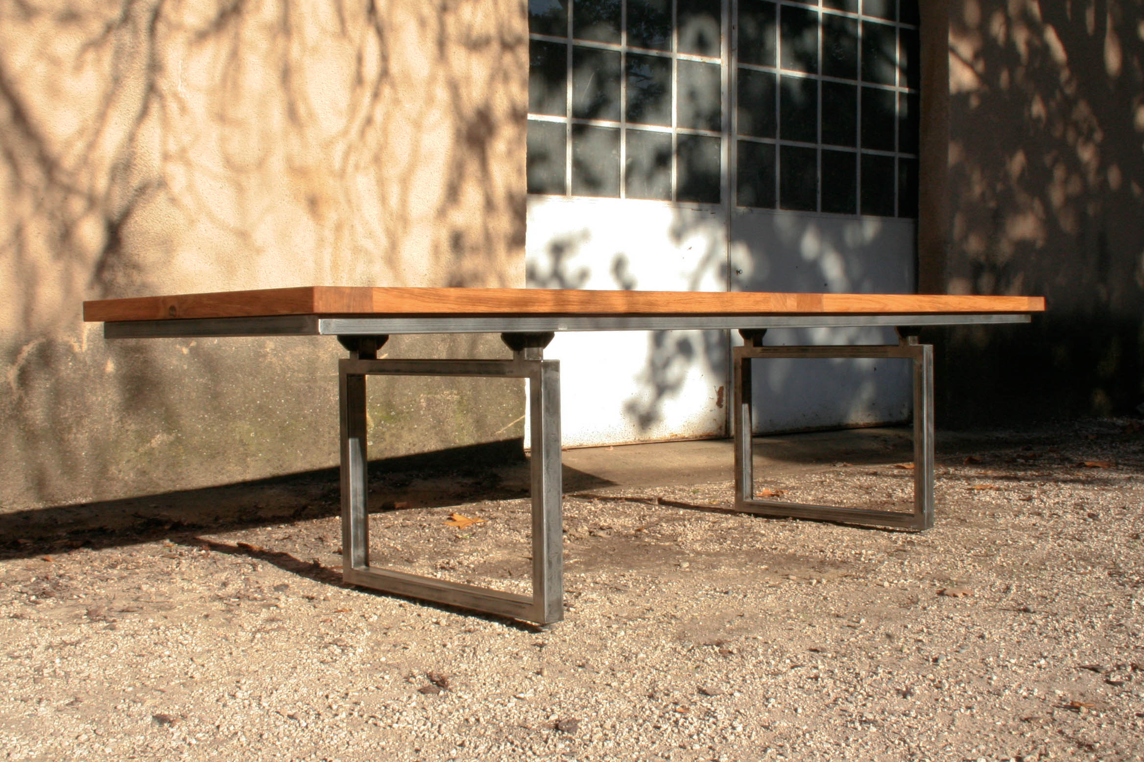 Table en bois avec un plateau rectangulaire soutenu par deux pieds en métal carrés, posée sur un sol gravillonné devant une porte vitrée.