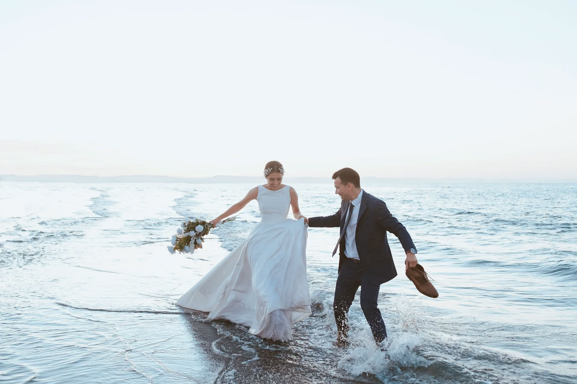 Mariée et marié riant pieds nus dans l'eau à la plage pendant une séance photo de mariage.