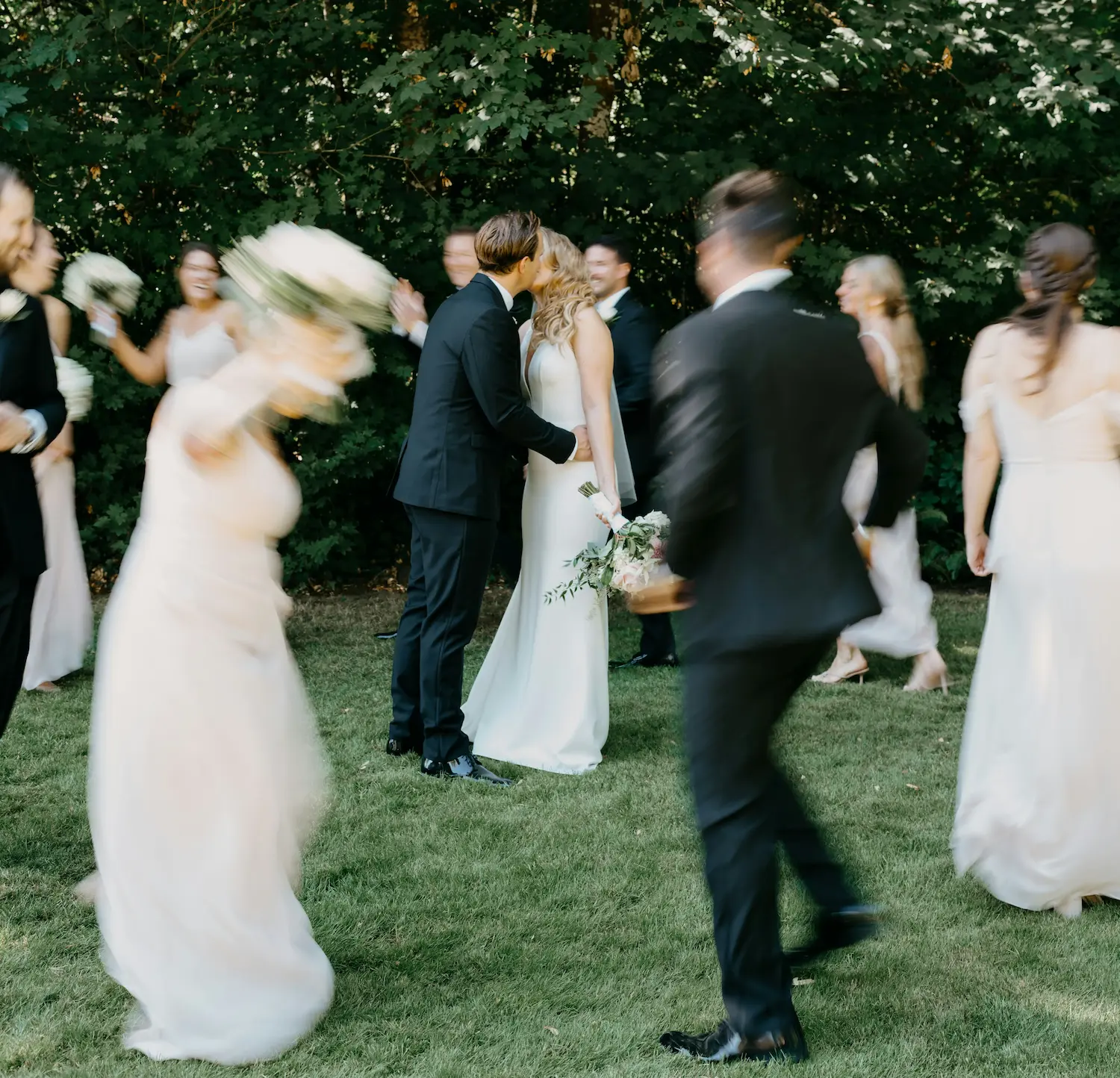 Un couple de mariés s'embrassant sur l'herbe entouré de leur cortège en mouvement flou.
