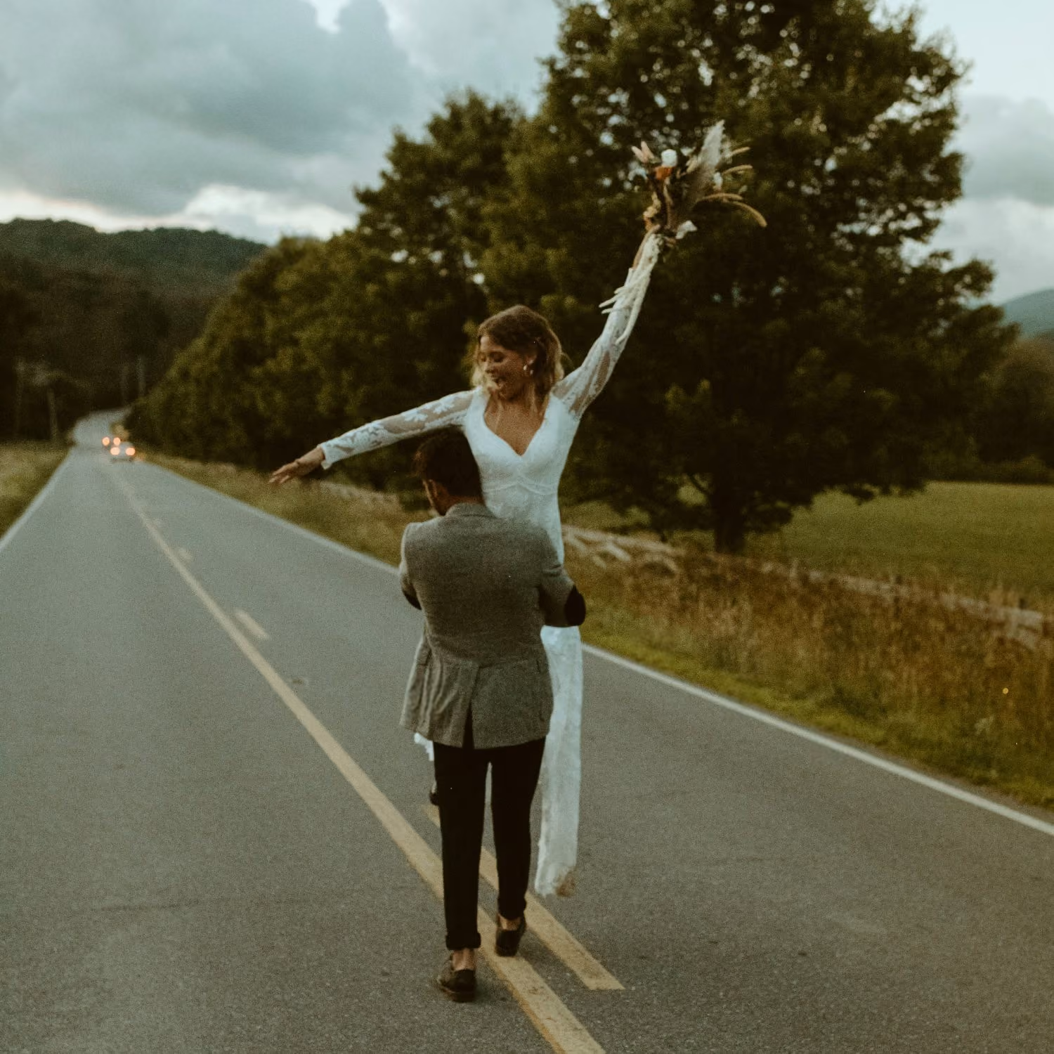 Un homme porte une mariée en robe blanche levant un bouquet sur une route bordée d'arbres.