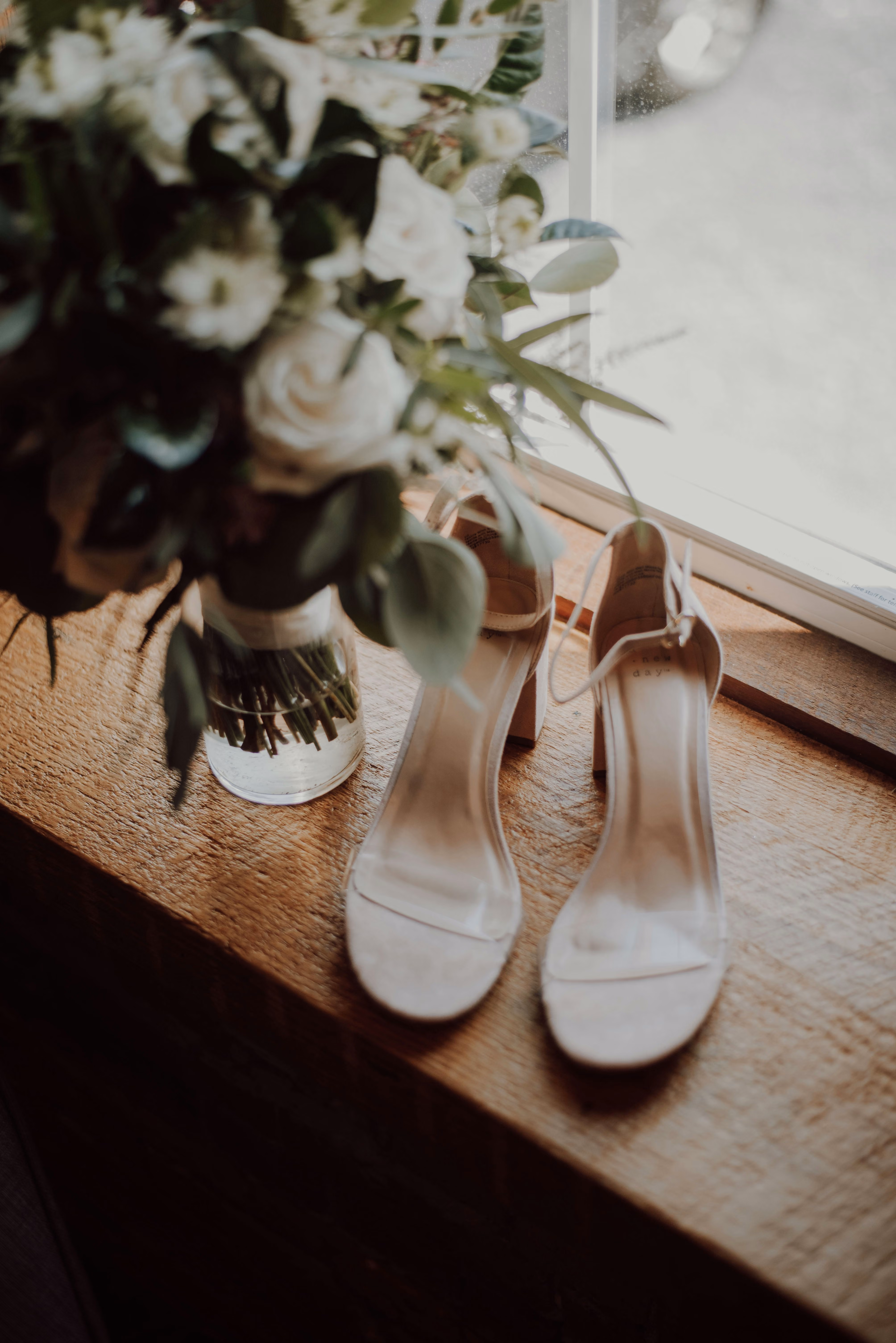 Chaussures à talons clairs près d'un bouquet de fleurs blanches sur un rebord de fenêtre en bois.