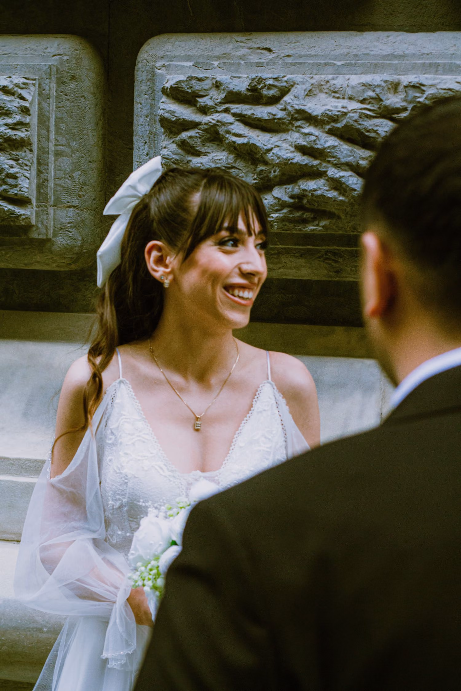 Mariée souriante en robe blanche avec bouquet regardant un homme en costume foncé de dos.
