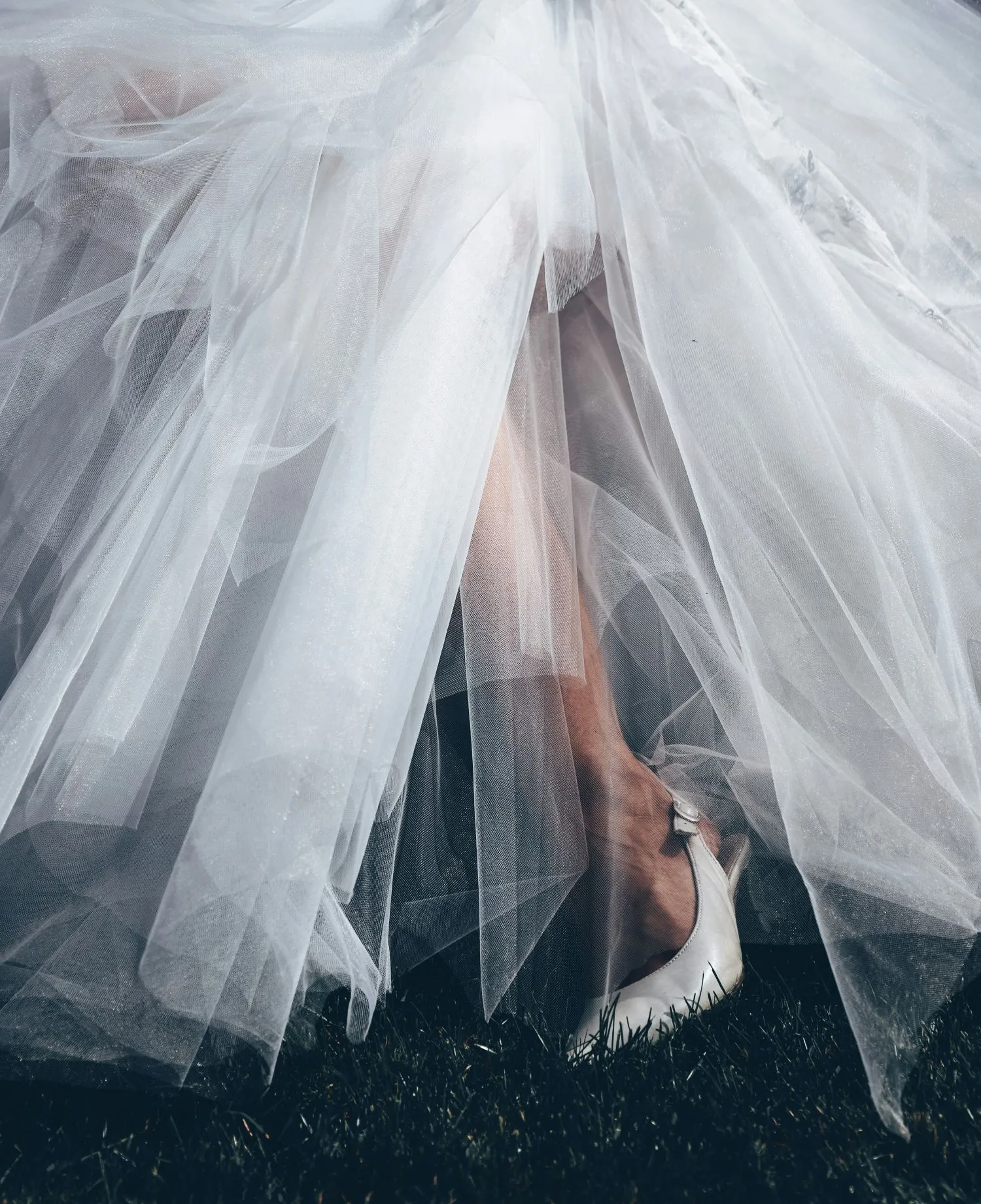 Un pied de mariée vêtue d'un chaussure blanche, entouré de tulle blanc, se dégageant sur l'herbe.