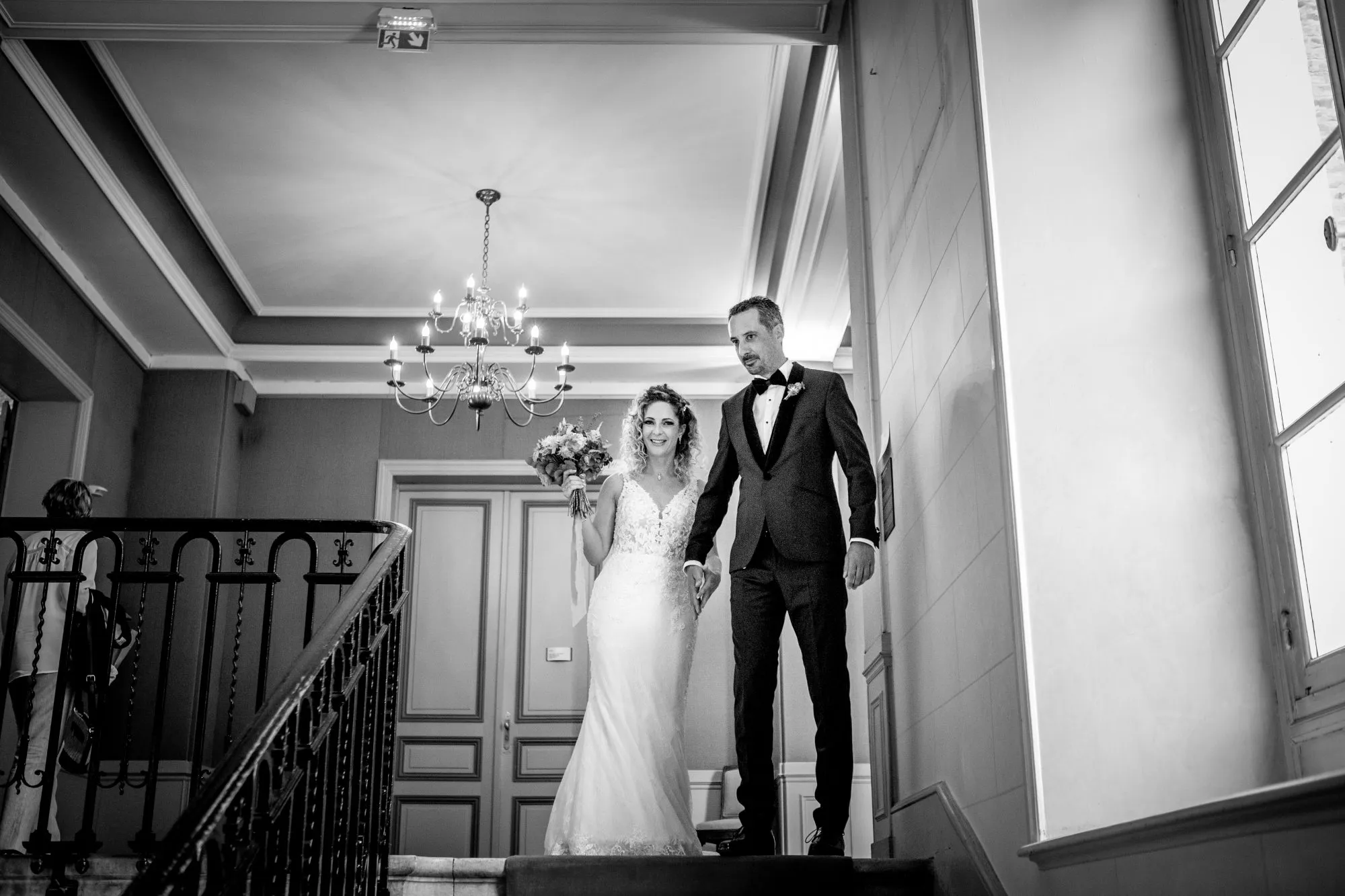 Une mariée rayonnante tient un bouquet de fleurs tandis qu'elle monte les escaliers avec son mari, dans un décor