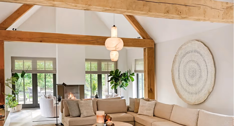 Lichte woonkamer met beige hoekbank, houten balken, vijgenplant en grote ronde wanddecoratie.