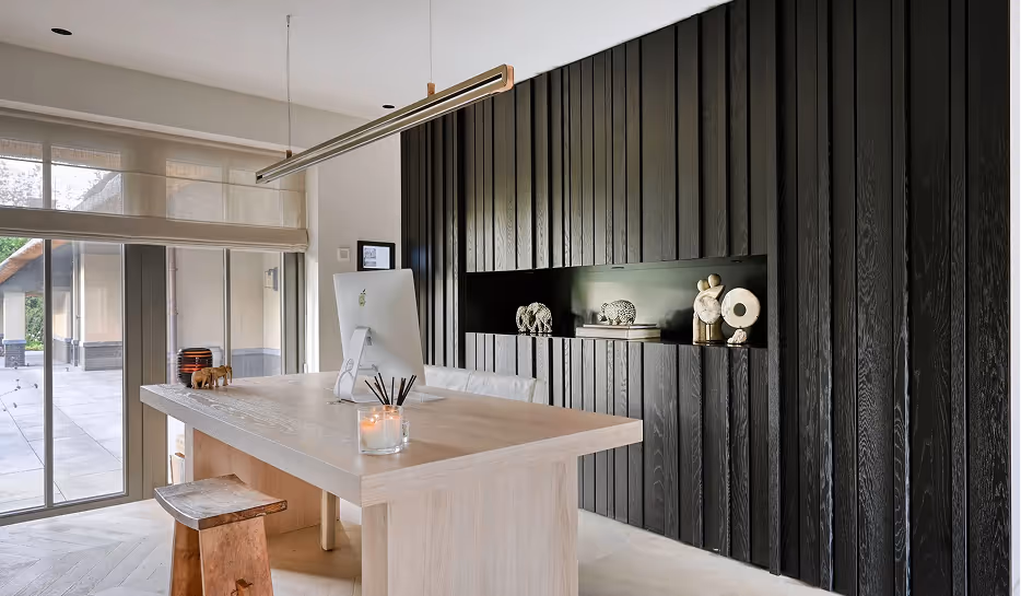 Moderne kantoorruimte met licht houten bureau, computer, houten kruk, en zwarte wand met decoraties.