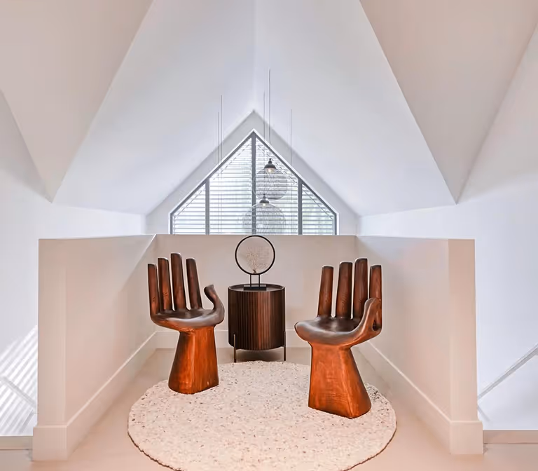 Twee handvormige houten stoelen op een rond wit tapijt in een lichte ruimte met een venster erachter.