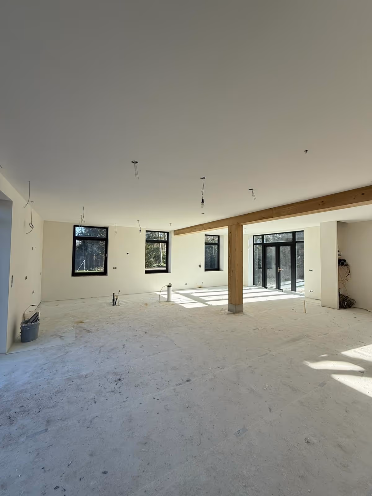 Grote lege kamer in aanbouw met witte muren, zwarte ramen en houten steunbalk in het midden.