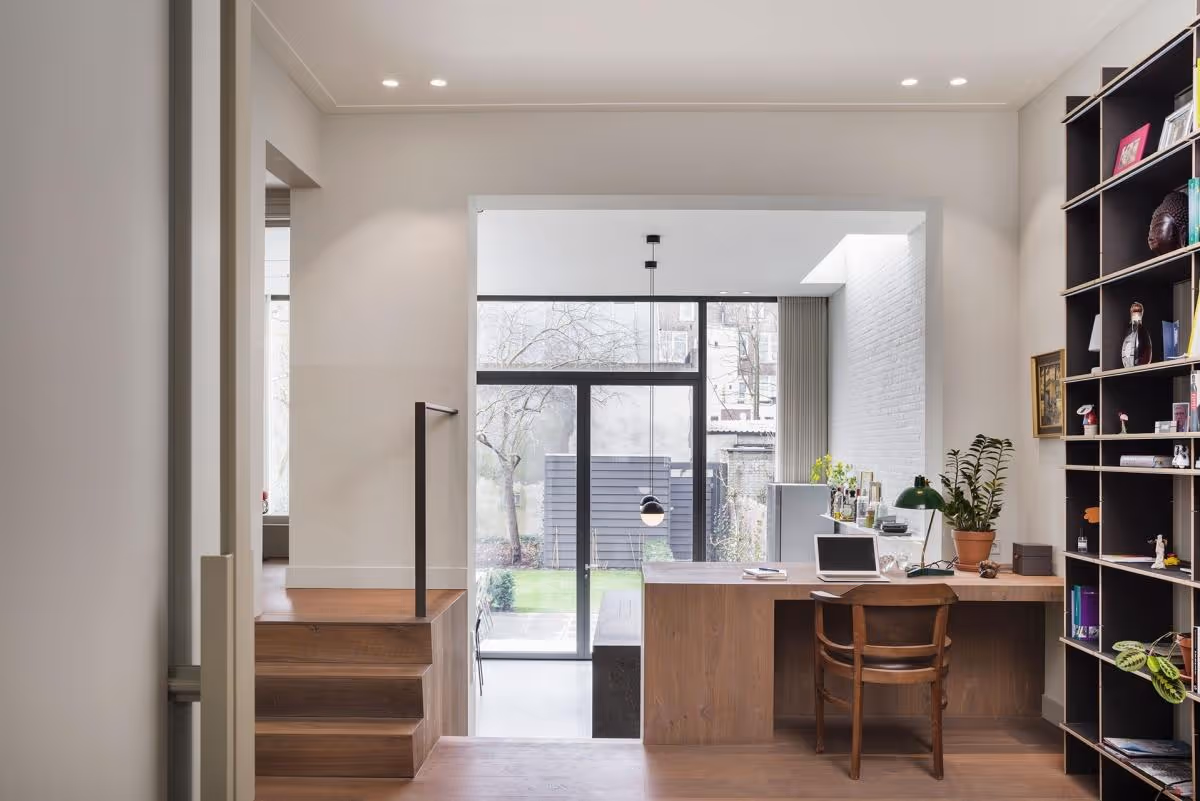 Modern thuiskantoor met houten bureau, stoel, laptop, planten en grote glazen schuifdeuren naar tuin.