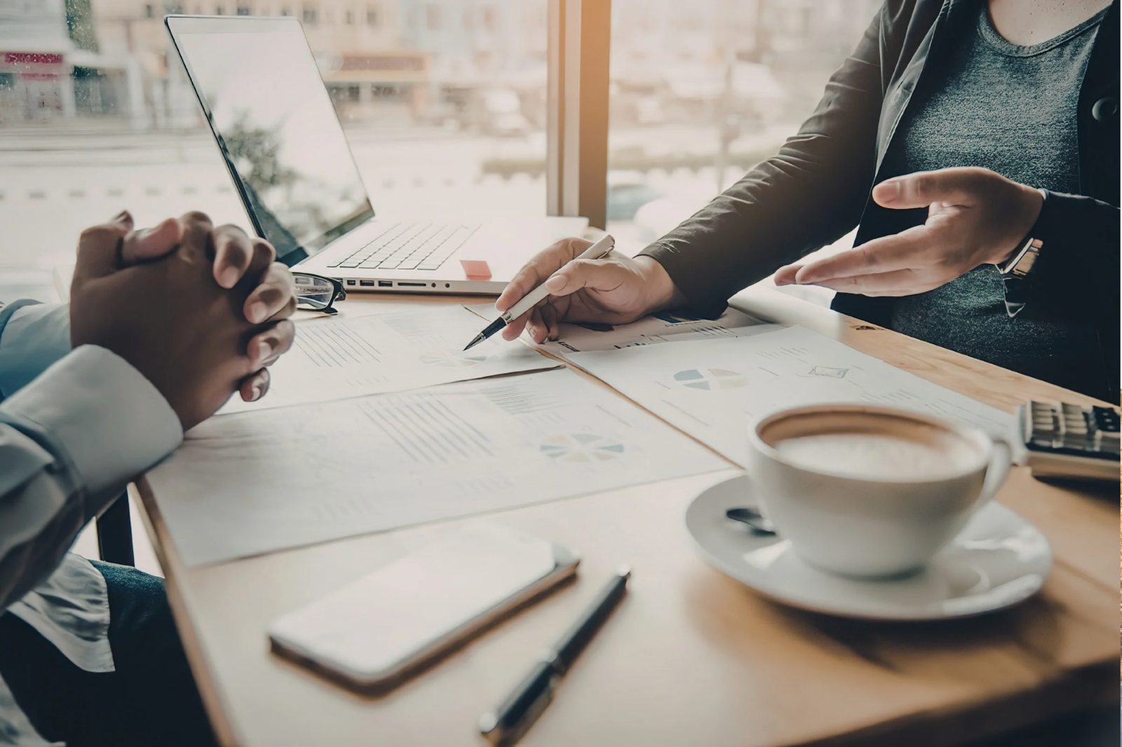 Two people discussing documents with charts at a table with laptop, coffee cup, and phone.