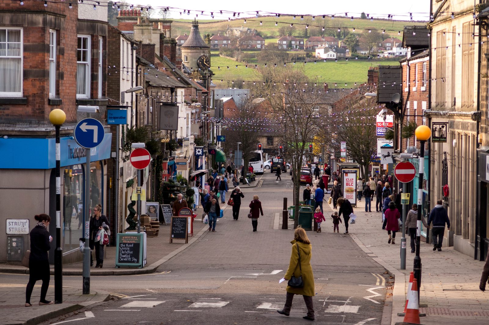 Busy town street with shops, pedestrians, holiday lights, and green hills in the background.