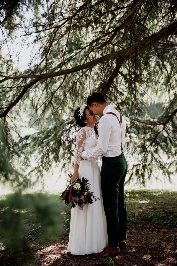 Jeunes mariés s'embrassant sous un grand arbre, elle en robe blanche dentelle et lui en pantalon vert et bretelles.