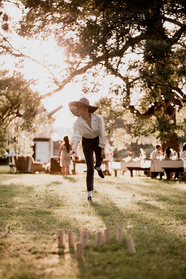 Une femme au chapeau joue au jeu de quilles lors d'un mariage bohème en plein air sous une lumière dorée.
