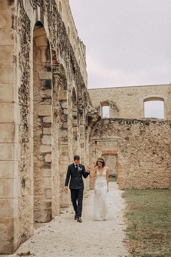 Jeunes mariés en tenue bohème chic se tenant la main dans les ruines en pierre d'un ancien bâtiment.