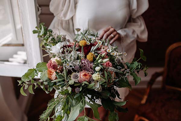 Mariée en robe blanche tenant un bouquet bohème aux tons rouge, rose et crème avec un feuillage sauvage.