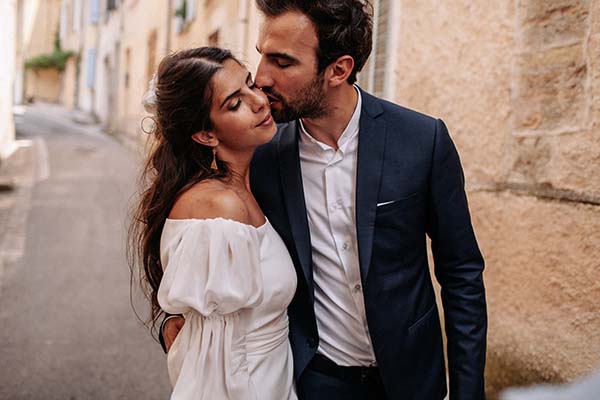 Jeune couple marié s'embrassant tendrement dans une ruelle ancienne, robe blanche bohème et costume bleu marine.