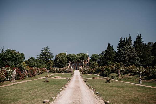 Allée de gravier blanc menant à un château entouré de jardins verdoyants sous un ciel bleu clair.