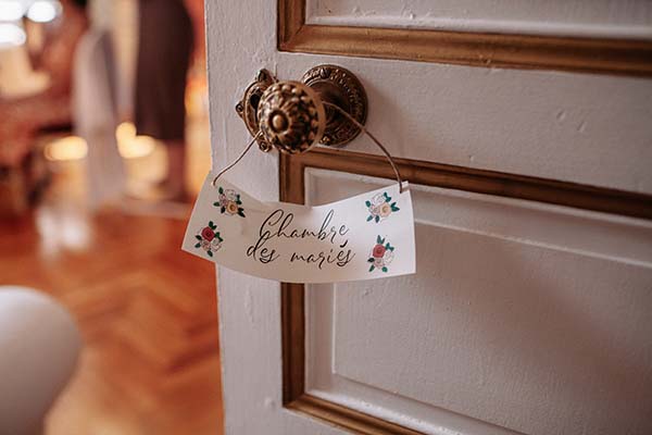 Plaque nominative fleurie « Chambre des mariés » suspendue à une poignée de porte blanche et dorée.