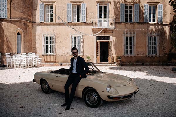 Homme élégant en costume noir et lunettes de soleil posant contre une décapotable beige devant un domaine ancien.