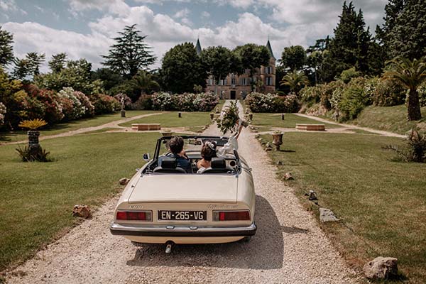 Jeunes mariés dans une décapotable crème sur un chemin de gravier menant à un château entouré de jardins luxuriants.