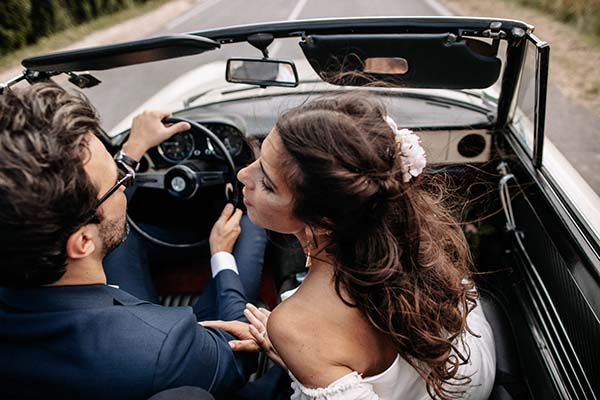 Jeunes mariés en tenue élégante s'embrassant dans une voiture décapotable vintage, ambiance romantique et bohème.