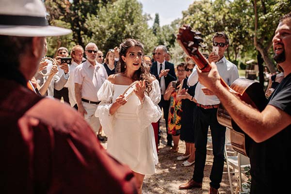 Une mariée en robe blanche rayonnante entourée d'invités et de musiciens lors d'une célébration festive en extérieur.