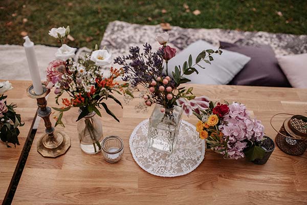 Décoration de table bohème avec bouquets de fleurs champêtres, bougies et coussins sur une table en bois clair.