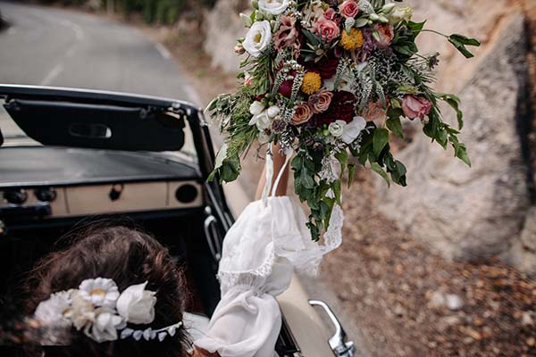 Une mariée en robe blanche tenant un bouquet bohème coloré, vue de dos dans une voiture décapotable.