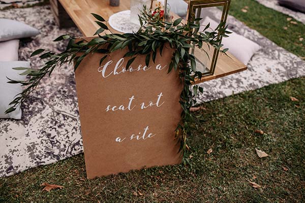 Panneau en carton kraft avec calligraphie blanche et feuillage vert pour un mariage bohème en plein air.