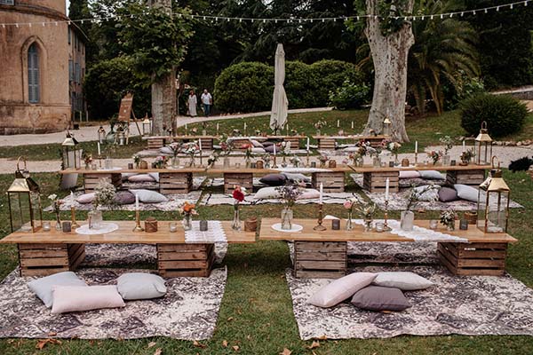Décoration de mariage bohème en plein air avec tables basses en bois, coussins et tapis sur une pelouse verdoyante.