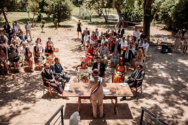 Cérémonie de mariage bohème en extérieur avec invités et officiant réunis sous des arbres dans un jardin ensoleillé.