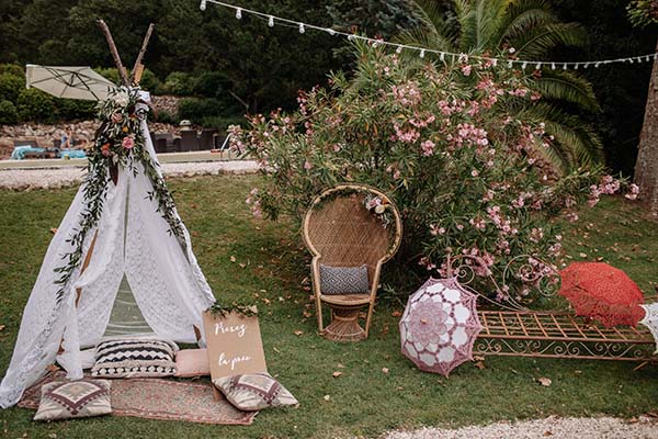 Espace détente bohème avec tipi blanc, fauteuil en rotin et fleurs roses dans un jardin luxuriant.